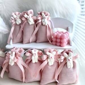 May include: Pink burlap favor bags with white bunny appliques and pink ribbon bows. The bags are arranged on a white shelf with a pink bubble candle.