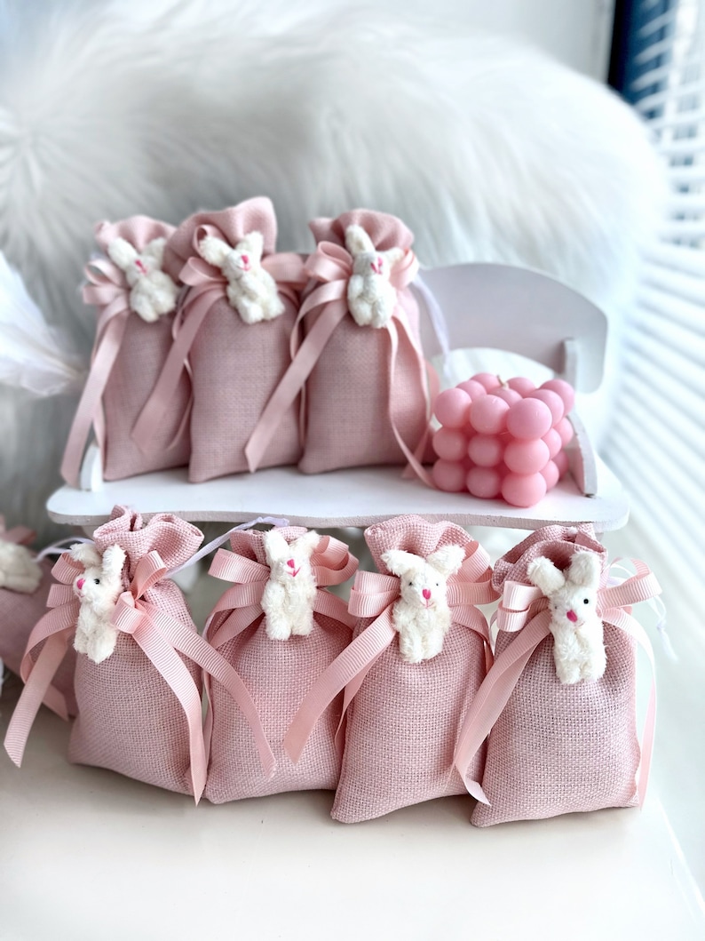 May include: Pink burlap favor bags with white bunny appliques and pink ribbon bows. The bags are arranged on a white shelf with a pink bubble candle.