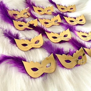 Puede incluir: M&aacute;scaras doradas de carnaval con plumas moradas. Cada m&aacute;scara tiene el texto "Jeane's Fifty & Fabulous" grabado.