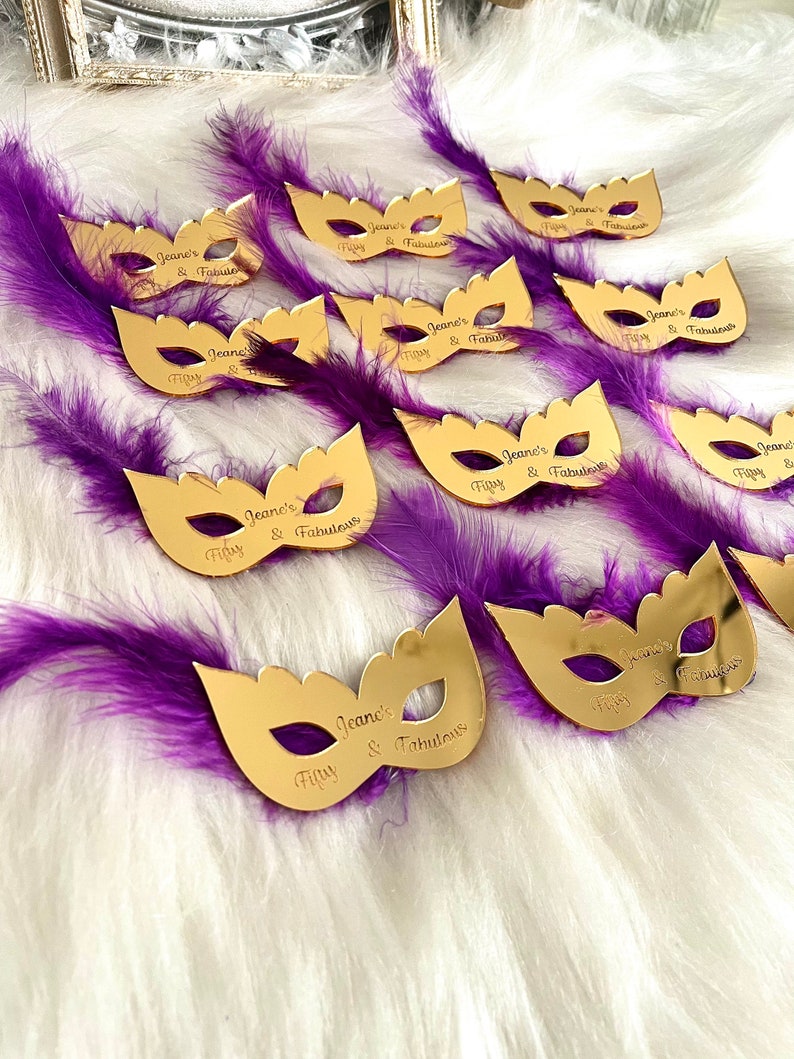 Puede incluir: M&aacute;scaras doradas de carnaval con plumas moradas. Cada m&aacute;scara tiene el texto "Jeane's Fifty & Fabulous" grabado.