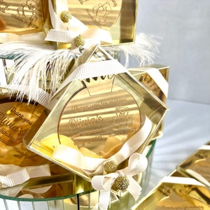 May include: Gold favor boxes with clear lids and white ribbon bows. Each box contains a round, gold mirror with the text "Thank you for making Olivia's first birthday very special" and a teddy bear graphic. The date "March 30, 2022" is also included.
