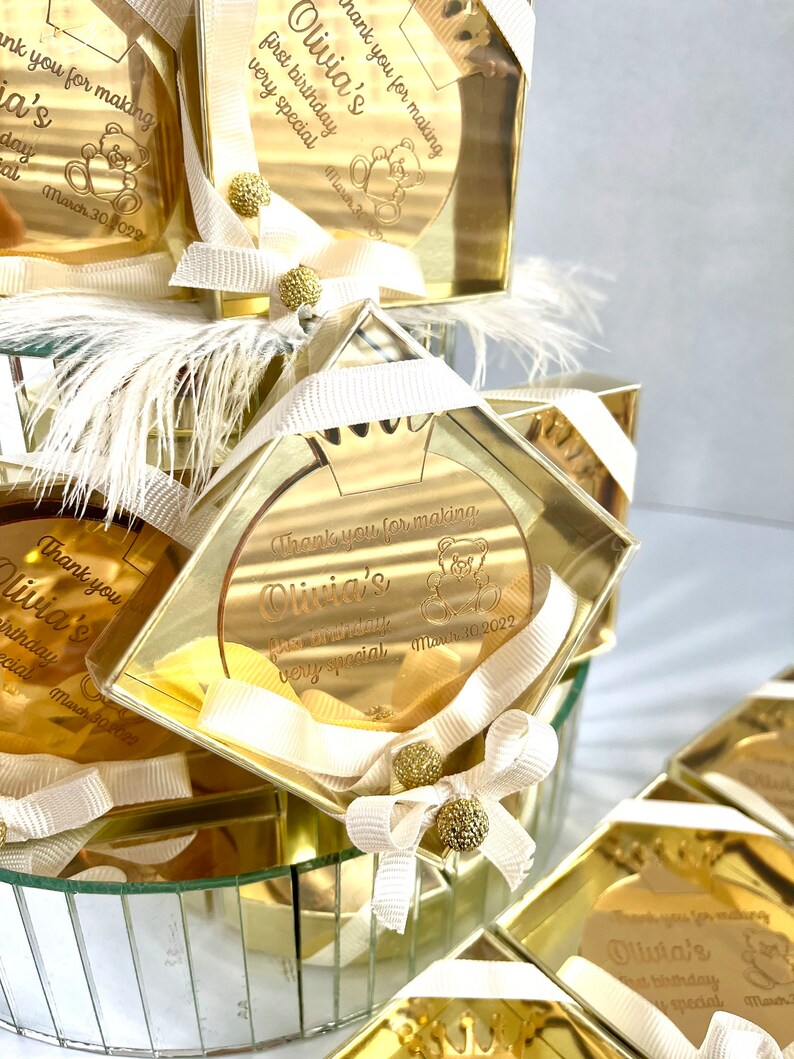 May include: Gold favor boxes with clear lids and white ribbon bows. Each box contains a round, gold mirror with the text "Thank you for making Olivia's first birthday very special" and a teddy bear graphic. The date "March 30, 2022" is also included.