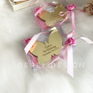 Puede incluir: Una peque&ntilde;a caja transparente con una cinta rosa y un espejo dorado en forma de mariposa en su interior. El espejo tiene la inscripci&oacute;n "Faith's 1st Birthday November 21st, 2020" grabada.