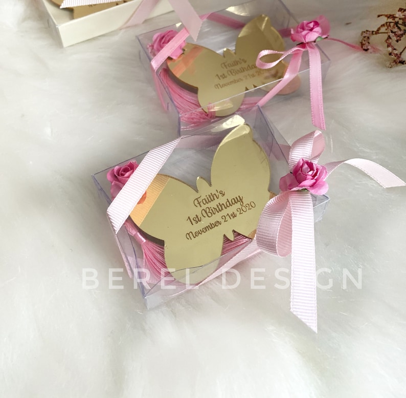 Puede incluir: Una peque&ntilde;a caja transparente con una cinta rosa y un espejo dorado en forma de mariposa en su interior. El espejo tiene la inscripci&oacute;n "Faith's 1st Birthday November 21st, 2020" grabada.