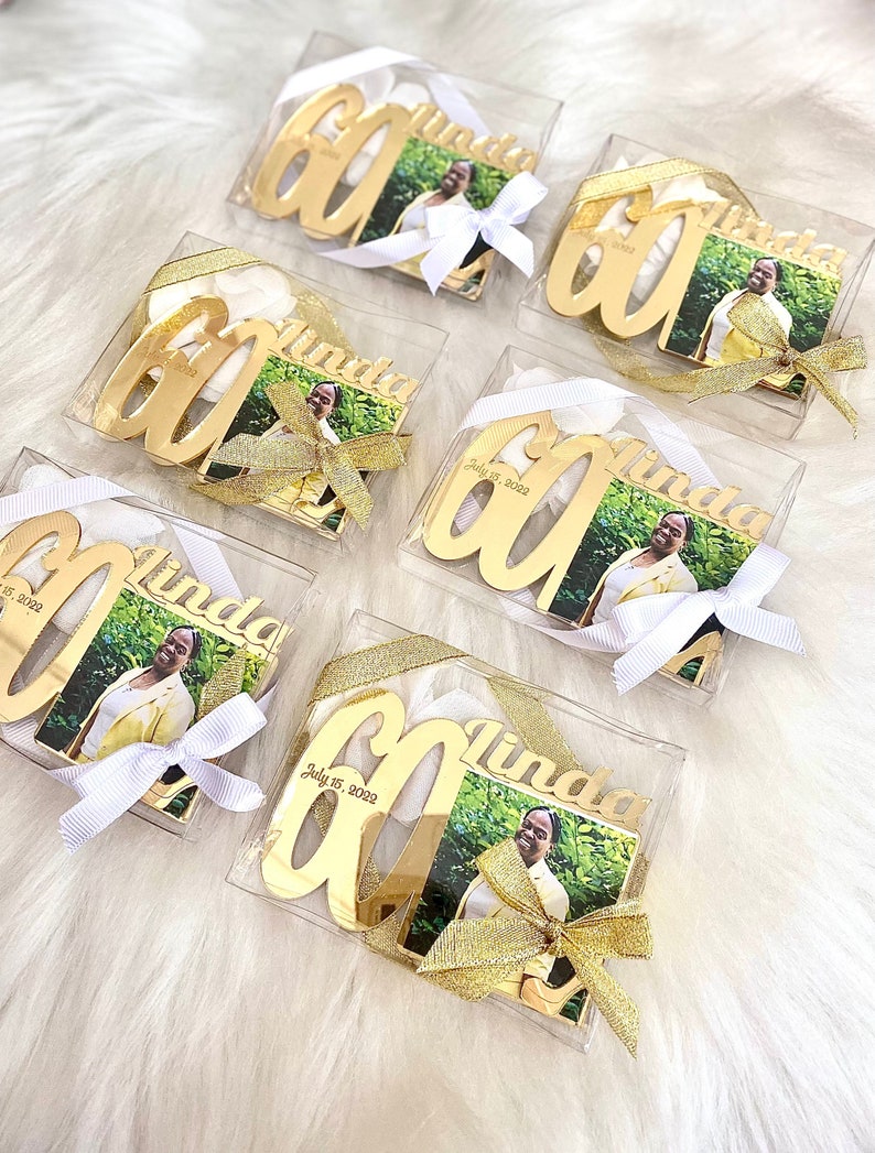 May include: Six clear plastic boxes with gold-colored metal ornaments shaped like the number 60. Each ornament has a photo of a person and the name "Linda" engraved on it. Some boxes also have the date "July 15, 2022" engraved on the ornament.