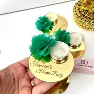 May include: Gold mirror candle holder with a green flower and a white tea light candle. The holder is engraved with the text "Tasneem's Henna Party".