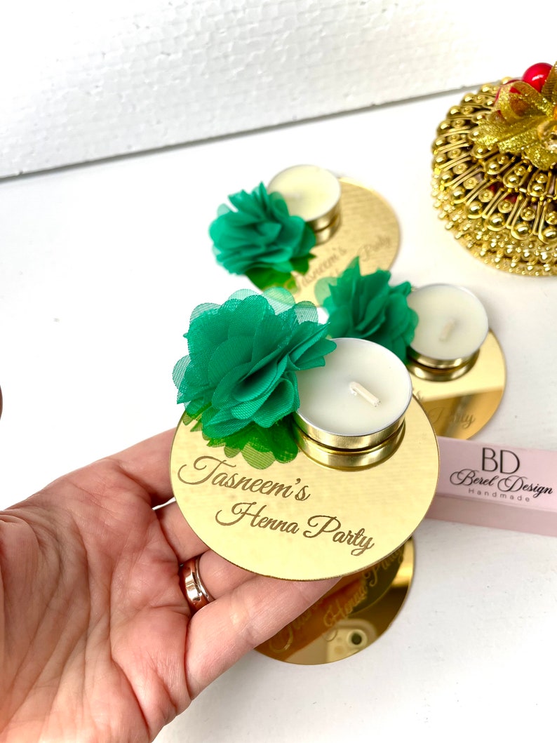 May include: Gold mirror candle holder with a green flower and a white tea light candle. The holder is engraved with the text "Tasneem's Henna Party".