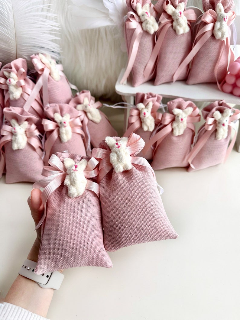 May include: Pink burlap favor bags with white bunny appliques and pink satin ribbon ties. The bags are arranged in a display on a white shelf.