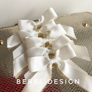 May include: Five white satin ribbon bows tied together in a cluster. The bows are arranged in a staggered pattern, with the top bow being the largest and the bottom bow being the smallest. The bows are on a gold background with a textured pattern.