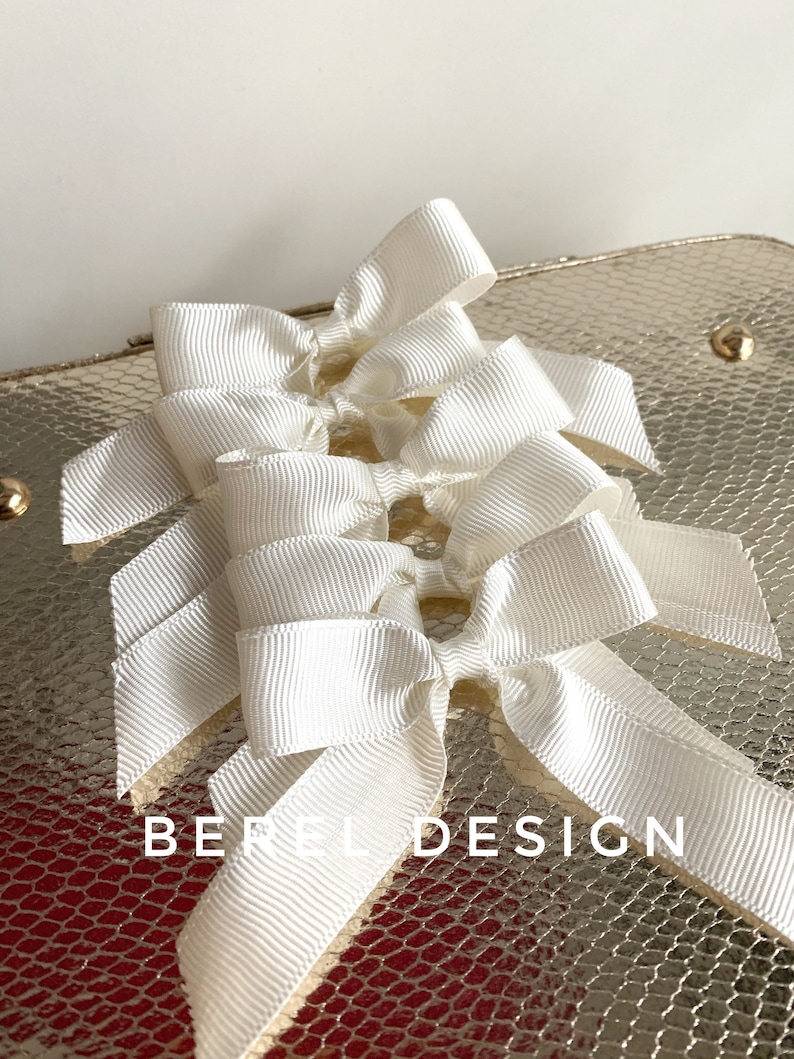 May include: Five white satin ribbon bows tied together in a cluster. The bows are arranged in a staggered pattern, with the top bow being the largest and the bottom bow being the smallest. The bows are on a gold background with a textured pattern.