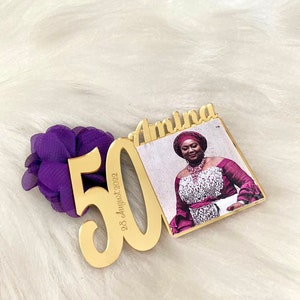 May include: Gold-colored acrylic plaque with the number 50 and the date "28 August 2022" inscribed. The plaque is attached to a smaller gold-colored acrylic frame with a photo of a woman wearing a purple and white dress and a head wrap. The frame has the name "Amina" inscribed on it.