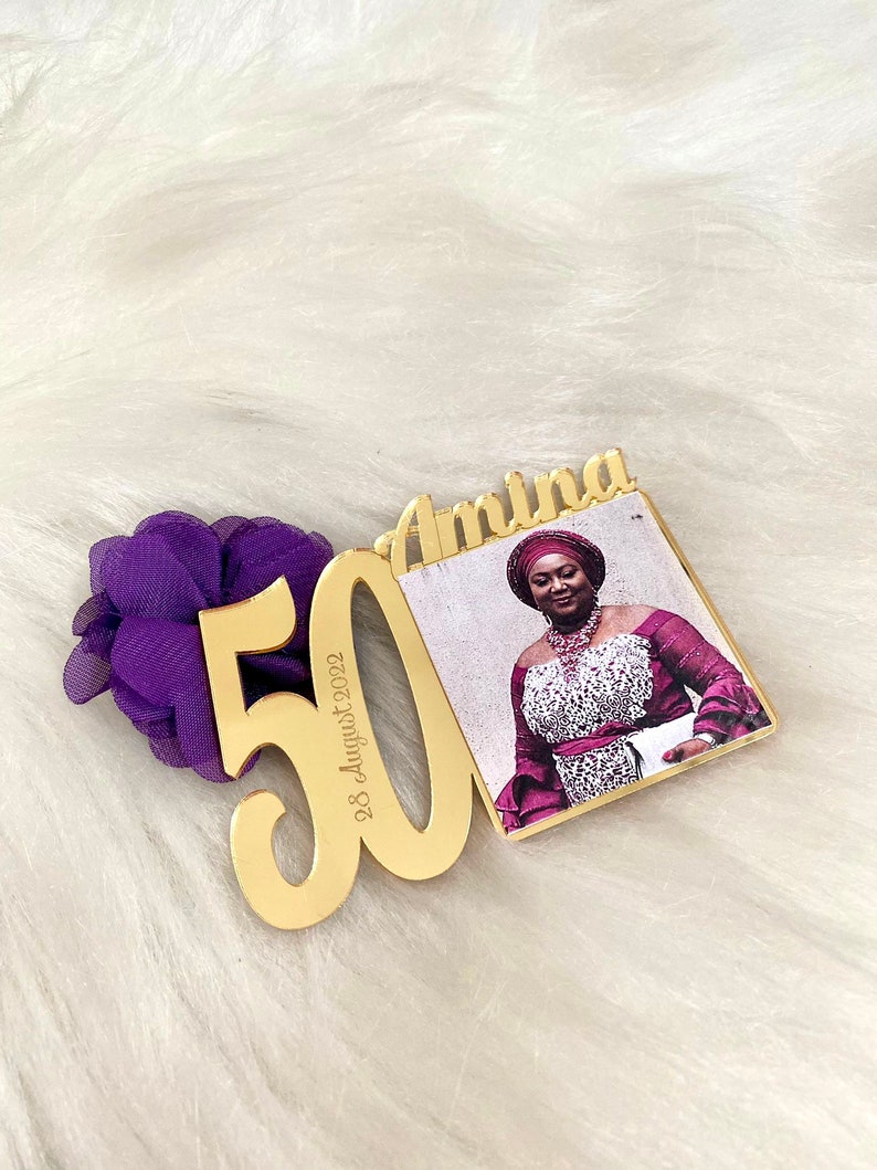 May include: Gold-colored acrylic plaque with the number 50 and the date "28 August 2022" inscribed. The plaque is attached to a smaller gold-colored acrylic frame with a photo of a woman wearing a purple and white dress and a head wrap. The frame has the name "Amina" inscribed on it.