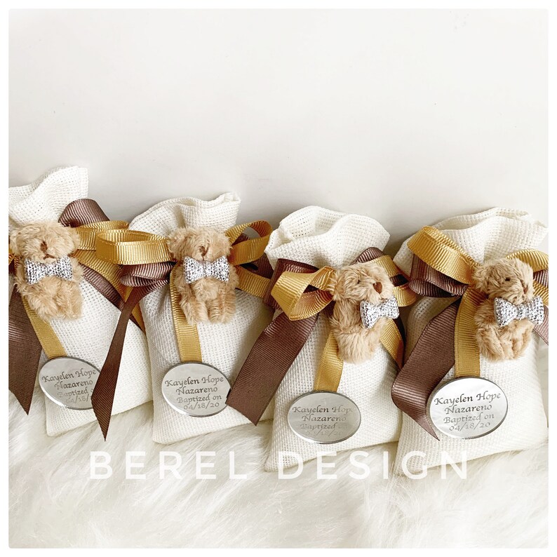 May include: Four white fabric gift bags with brown and gold ribbon bows. Each bag has a small plush teddy bear with a bow tie and a silver oval tag with the text "Kayelen Hope Nazareno Baptized on 04/18/20".