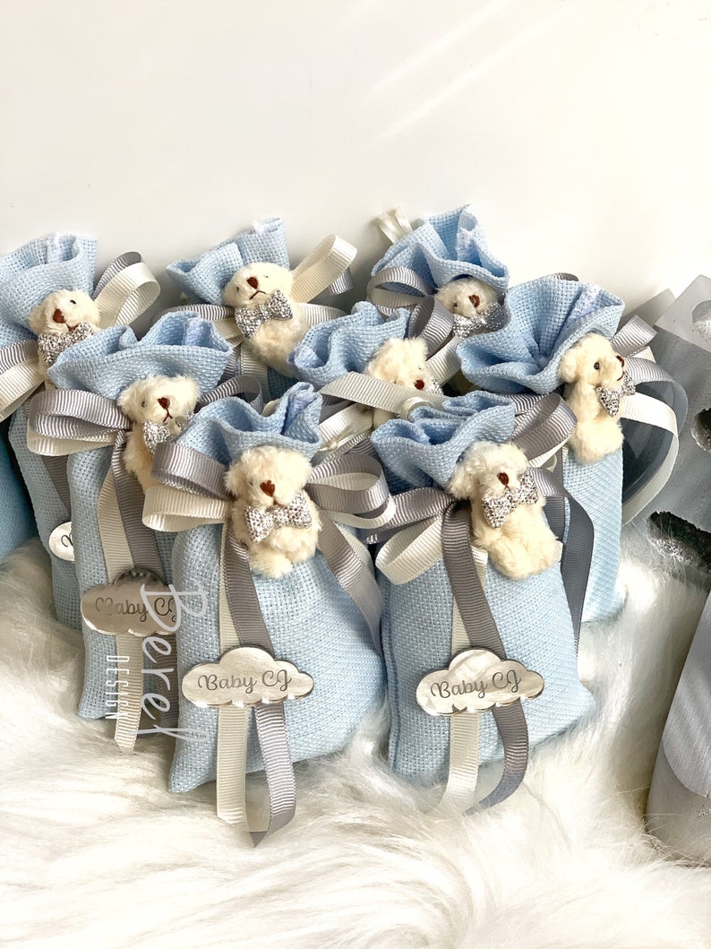 Puede incluir: Bolsas de tela azul con ositos de peluche blancos y lazos plateados. Cada bolsa tiene una etiqueta plateada con el texto "Baby CJ" en ella.