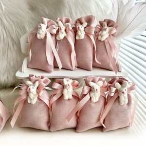 May include: Pink burlap favor bags with white bunny rabbit embellishments and pink satin ribbon ties. The bags are arranged on a white wooden surface.