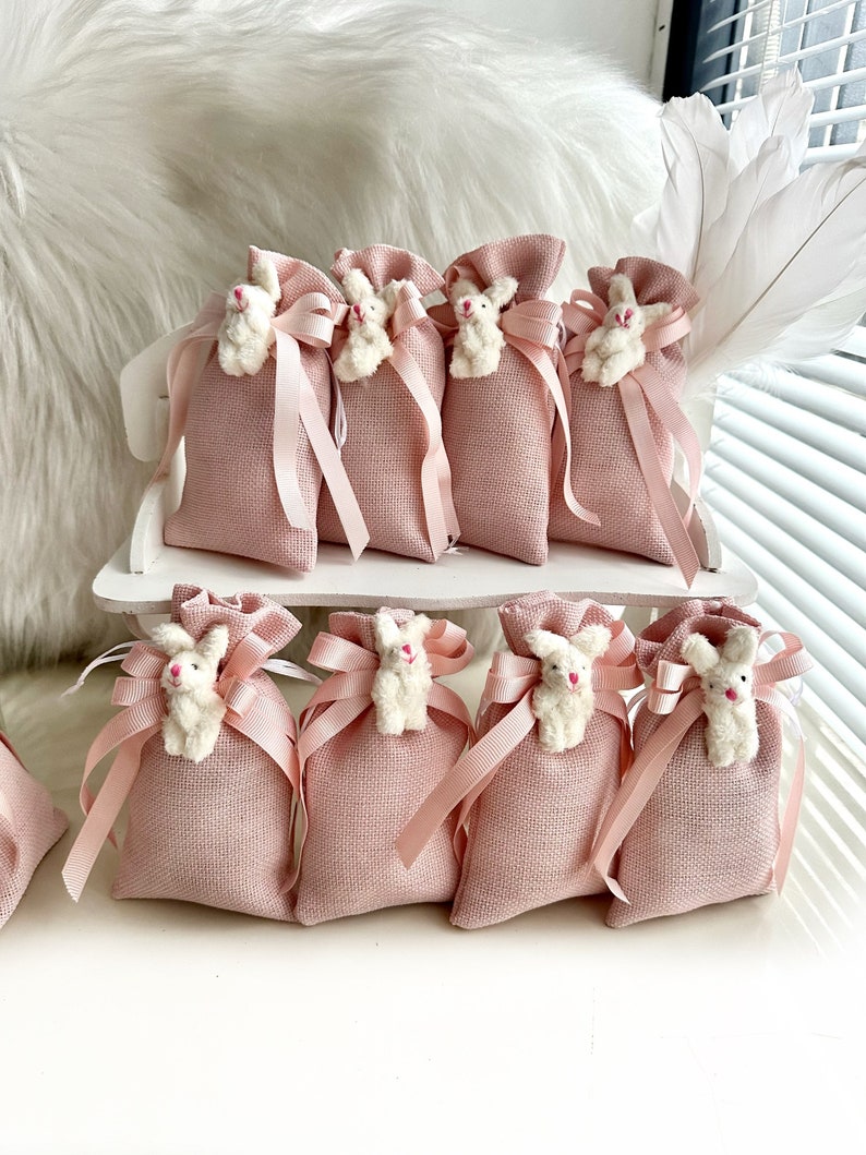 May include: Pink burlap favor bags with white bunny rabbit embellishments and pink satin ribbon ties. The bags are arranged on a white wooden surface.