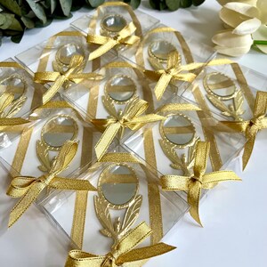May include: A collection of gold-toned handheld mirrors, each encased in a clear square box. Every mirror features a decorative handle and is adorned with a shimmering gold ribbon tied in a bow around the exterior of the transparent packaging.