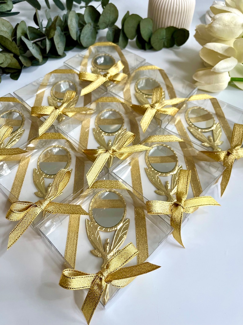 May include: A collection of gold-toned handheld mirrors, each encased in a clear square box. Every mirror features a decorative handle and is adorned with a shimmering gold ribbon tied in a bow around the exterior of the transparent packaging.