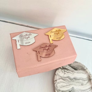 May include: Three gold, silver, and rose gold graduation caps with the text "Smallville High 2024" on a pink box.