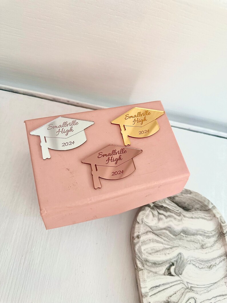 May include: Three gold, silver, and rose gold graduation caps with the text "Smallville High 2024" on a pink box.