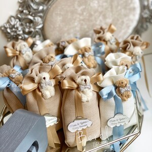 May include: A collection of small, beige fabric bags with a teddy bear charm and a blue ribbon. Each bag has a tag with the text "Mari & Yanis's Baby Shower".