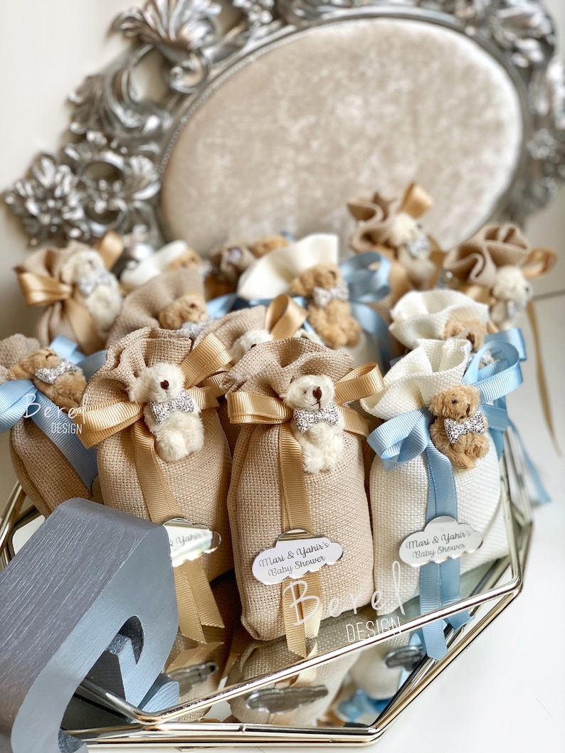 May include: A collection of small, beige fabric bags with a teddy bear charm and a blue ribbon. Each bag has a tag with the text "Mari & Yanis's Baby Shower".