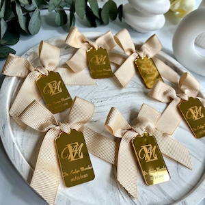 May include: Gold favor tags with a cream-colored satin ribbon bow. The tags are engraved with the letter "E" and the text "Dr. Evelyn Mancha 05/15/2023".