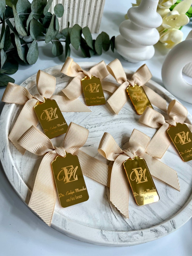 May include: Gold favor tags with a cream-colored satin ribbon bow. The tags are engraved with the letter "E" and the text "Dr. Evelyn Mancha 05/15/2023".