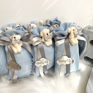 Puede incluir: Bolsas de regalo azul claro con un peque&ntilde;o osito de peluche blanco que lleva una pajarita. Cada bolsa tiene una etiqueta en forma de nube plateada con el texto "Baby CJ" en ella.