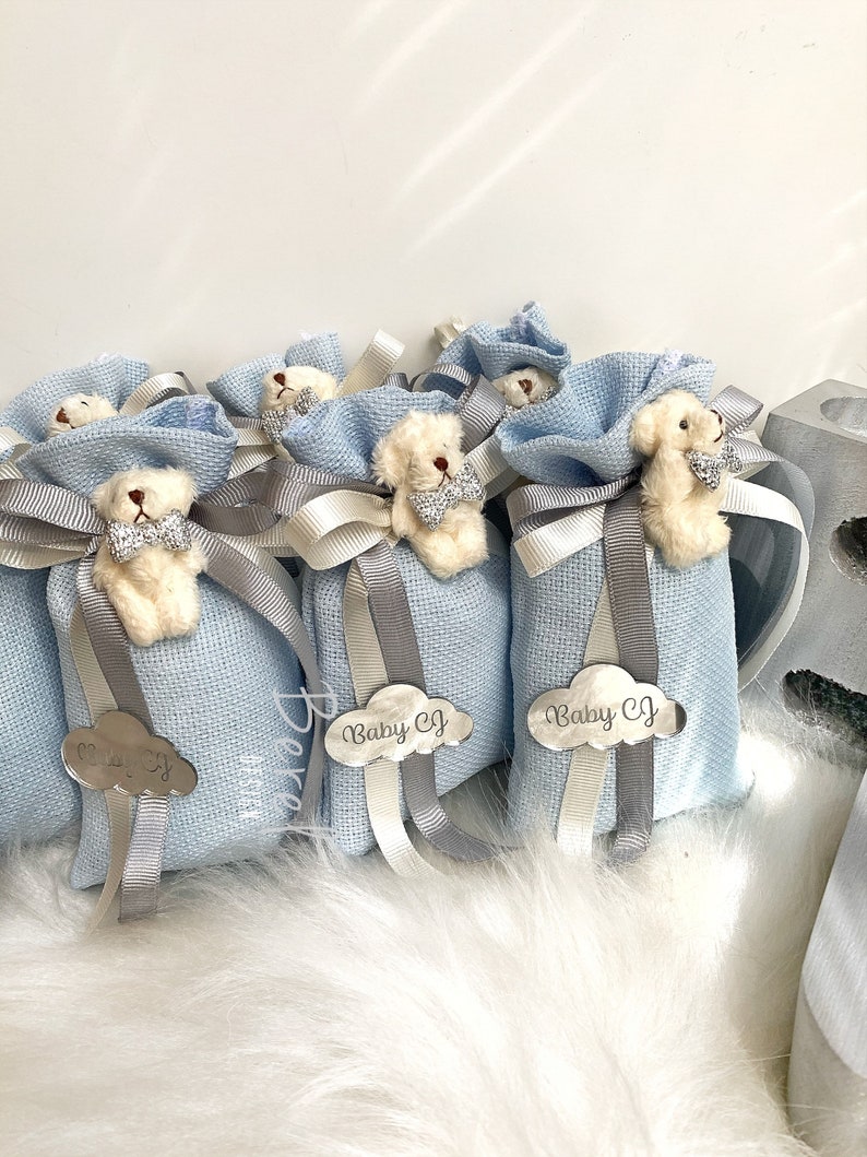 Puede incluir: Bolsas de regalo azul claro con un peque&ntilde;o osito de peluche blanco que lleva una pajarita. Cada bolsa tiene una etiqueta en forma de nube plateada con el texto "Baby CJ" en ella.