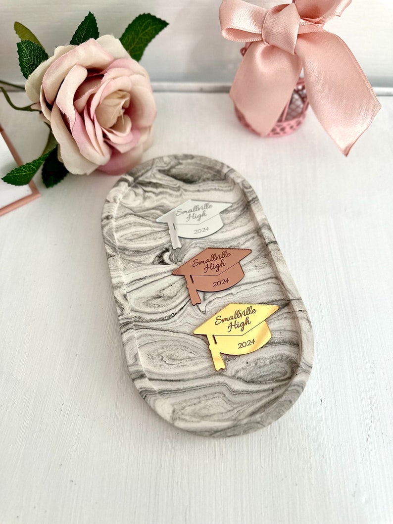 May include: Three graduation caps with the text "Smallville High 2024" on a marble tray. The caps are silver, rose gold, and gold.