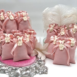 May include: Pink fabric favor bags with white bunny appliques and pink ribbon ties. The bags are arranged on a silver tray with a pink velvet lining.
