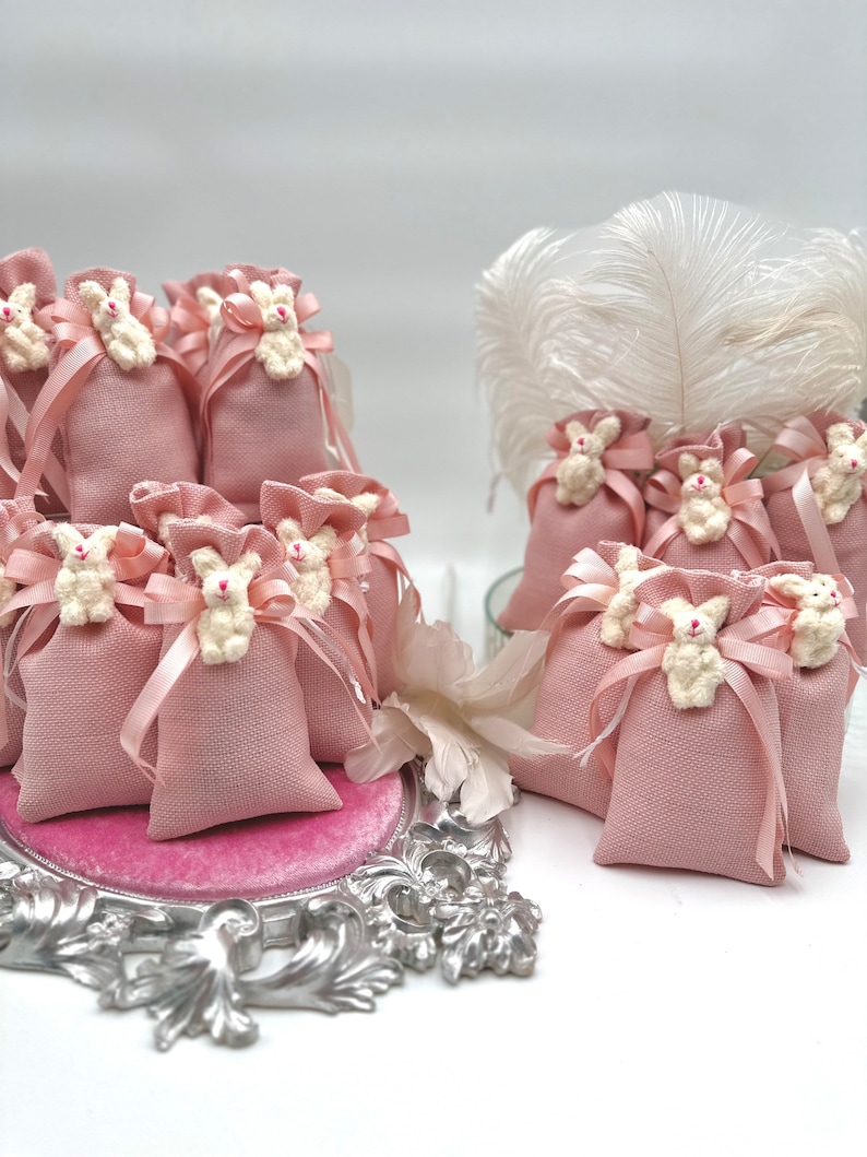 May include: Pink fabric favor bags with white bunny appliques and pink ribbon ties. The bags are arranged on a silver tray with a pink velvet lining.