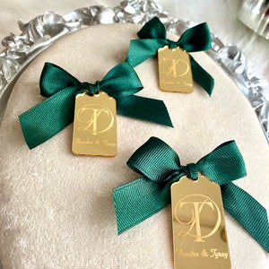 May include: Three gold mirror tags with green bows. Each tag has a cursive letter "T" and the text "Sandra & Tray" engraved on the front.
