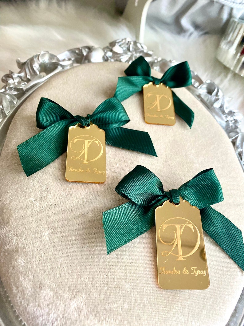 May include: Three gold mirror tags with green bows. Each tag has a cursive letter "T" and the text "Sandra & Tray" engraved on the front.
