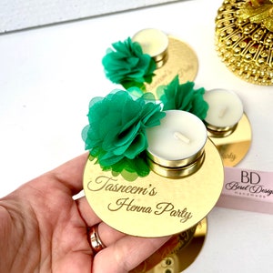 May include: Gold round mirror with the text "Tasneem's Henna Party" engraved on it. A small white tea light candle sits on top of the mirror, and a green fabric flower is placed on the candle.