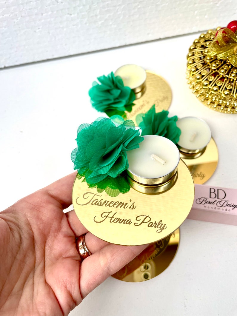 May include: Gold round mirror with the text "Tasneem's Henna Party" engraved on it. A small white tea light candle sits on top of the mirror, and a green fabric flower is placed on the candle.