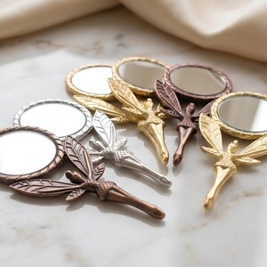 May include: A collection of five small handheld mirrors, each featuring a metallic handle shaped like a winged figure. The mirrors are finished in various metallic tones including gold, silver, copper, and bronze, with circular glass surfaces.