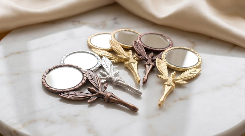 May include: A collection of five small handheld mirrors, each featuring a metallic handle shaped like a winged figure. The mirrors are finished in various metallic tones including gold, silver, copper, and bronze, with circular glass surfaces.