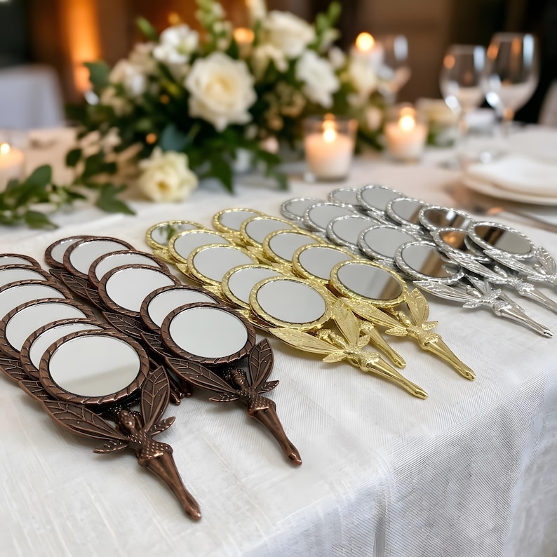 May include: A collection of round handheld mirrors with metallic finishes in bronze, gold, and silver. Each mirror features a handle shaped like a fairy with wings and a pointed tip. The mirrors are arranged in rows on a white tablecloth.