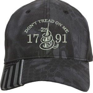 May include: A black camouflage baseball cap with a gray American flag on the side. The front of the cap features a green embroidered snake with the text "Don't Tread On Me" and "1776" above and "91" below.
