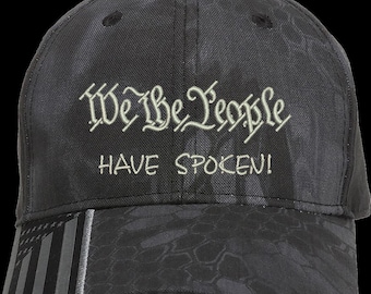 以下が含まれることがあります： 「We the People Have Spoken!」と白く刺繍されたデザインが施された、黒の迷彩柄の野球帽。帽子には、側面に白黒のアメリカ国旗がデザインされています。
