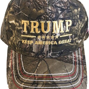 May include: A camouflage baseball cap with the text "TRUMP KEEP AMERICA GREAT" embroidered in gold on the front. The cap has a red, white, and blue American flag patch on the back.