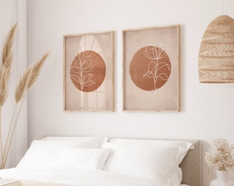 Puede incluir: Dos impresiones enmarcadas con un diseño minimalista que presenta un círculo marrón con un dibujo de línea blanca de una planta en su interior. Las impresiones están colgadas en una pared blanca sobre una cama blanca con una manta beige.