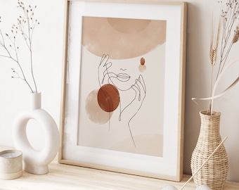 Op de afbeelding: Een minimalistische line art print van een vrouwen gezicht met een hoed, omringd door abstracte vormen in tinten bruin en oranje. De print is ingelijst in een licht houten lijst.