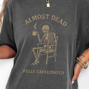 May include: A charcoal grey short-sleeve t-shirt featuring a graphic of a skeleton sitting in a chair holding a coffee mug. The text ALMOST DEAD is printed above the skeleton and FULLY CAFFEINATED is printed below in a light tan color.
