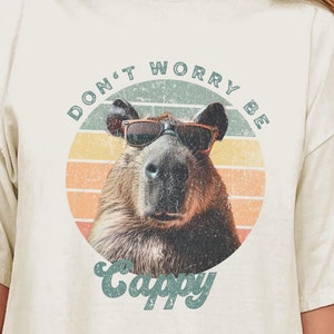 Woman wearing an ivory t-shirt with a retro graphic of a capybara in sunglasses. The text says 'Don't Worry Be Cappy.' Ideal for capybara fans, funny animal shirt lovers, and vintage tee enthusiasts. A cute gift for those who enjoy animal memes.