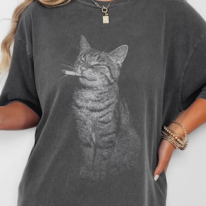 May include: Dark grey oversized t-shirt with a graphic of a cat smoking a cigarette. The cat is depicted with its eyes closed and a smile. The shirt has a loose fit and short sleeves. The text "PEPPER" and "99 ORGANIZERS" are at the bottom.
