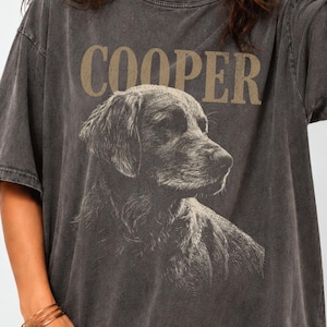May include: A charcoal grey oversized t-shirt featuring a graphic illustration of a dog in profile. The word COOPER is printed in large, tan, capital letters above the dog. The fabric has a faded, washed-out appearance with short sleeves.