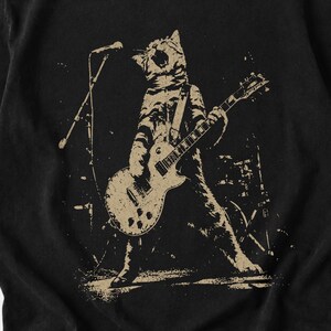 Punk cat design featuring a fierce tabby cat standing onstage while playing an electric guitar beneath a microphone. The distressed monochrome artwork captures the energy of a live rock performance with grunge texture, bold movement, and vintage concert poster style. A striking graphic for cat lovers, guitar players, alternative music fans, punk rock enthusiasts, and shoppers seeking edgy band-inspired apparel.
